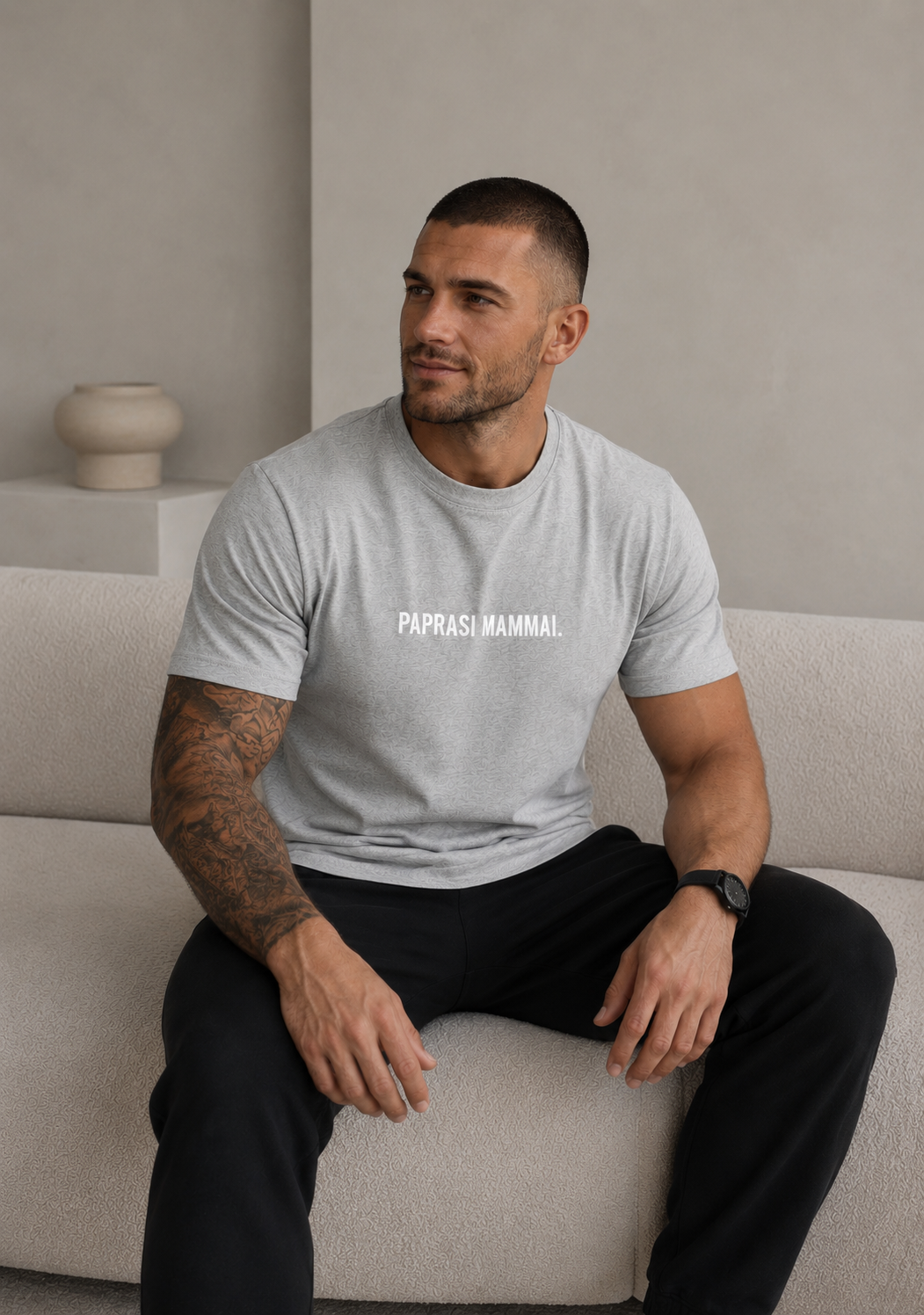 A man sitting on a light beige sofa wearing a light gray crew-neck t-shirt with 'PAPRASI MAMMAI' printed in white across the chest, paired with black pants.