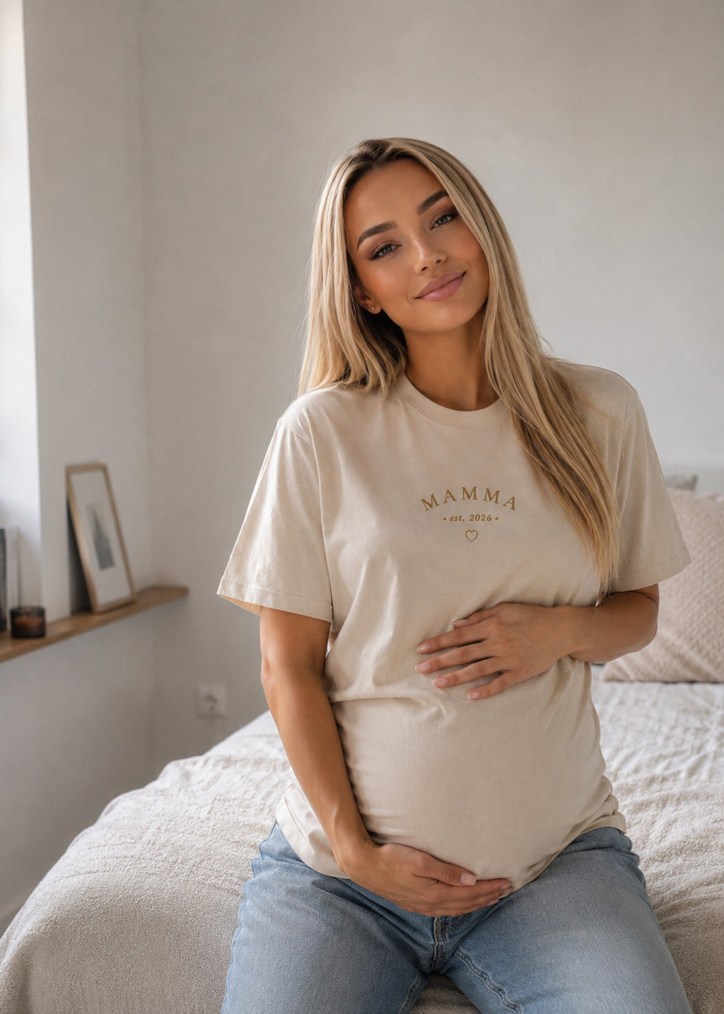 A pregnant woman with blonde hair sits on a bed wearing a beige t-shirt with 'MAMMA EST. 2021' printed in gold on the chest, paired with light blue jeans.