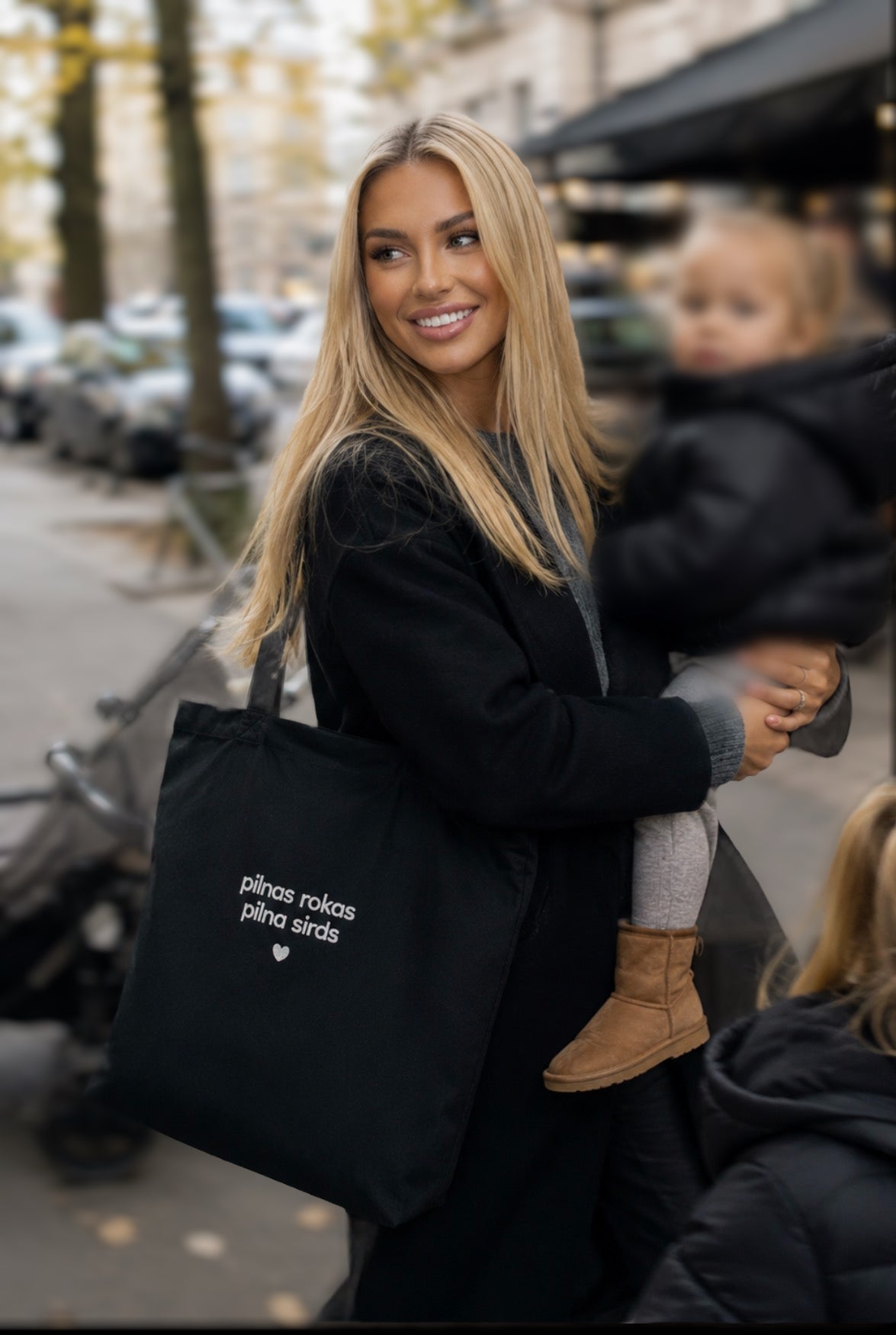 A woman carrying a black tote bag with white text reading 'pilnas rokas pilna sirds' on a city street, with a child in a black coat beside her.