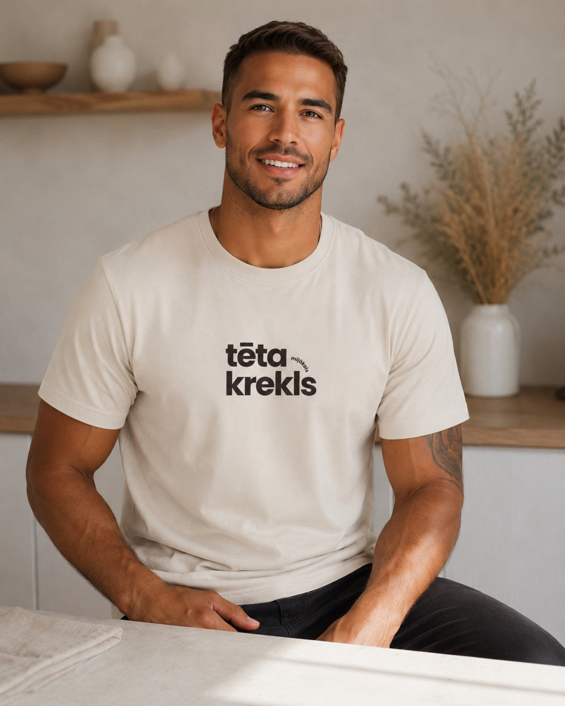 A smiling man wearing a cream-colored t-shirt with the black text 'tēta mīļākais krekls" printed on the chest, seated in a brightly lit, modern interior setting.