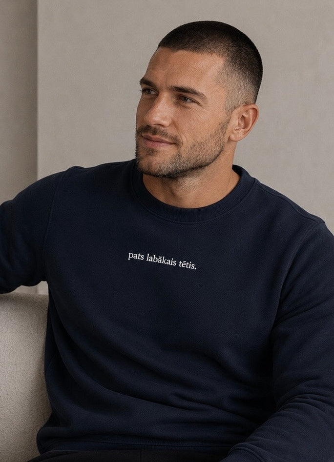 A man wearing a navy blue crewneck sweatshirt with white embroidered text on the chest, sitting on a light beige sofa in a minimalist room.