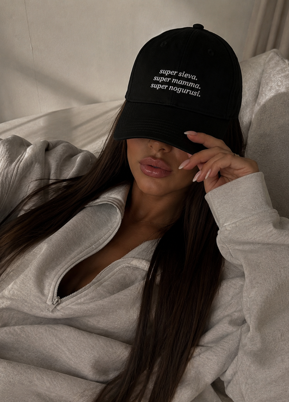 A woman wearing a black baseball cap with white embroidered text reading 'super sieva, super mamma, super nogurusi.' while lying on a bed in a light gray sweatshirt.