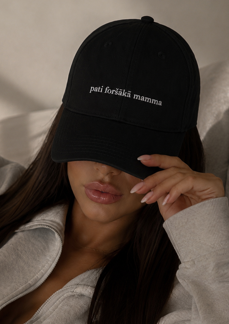 A woman wearing a black baseball cap with white embroidered text reading 'pati foršākā mamma' and a light gray zip-up hoodie, resting against a neutral background.