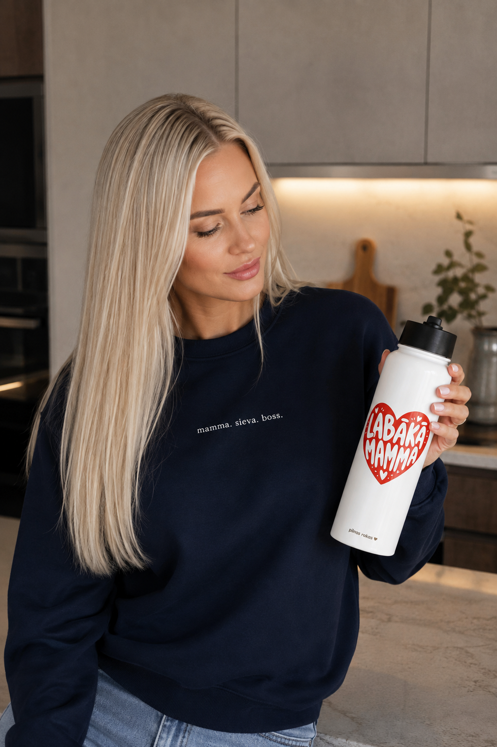 A blonde woman in a navy sweatshirt holds a white insulated water bottle with a black lid. The bottle has a red heart graphic with the words 'LABĀKĀ MAMMA' in white.
