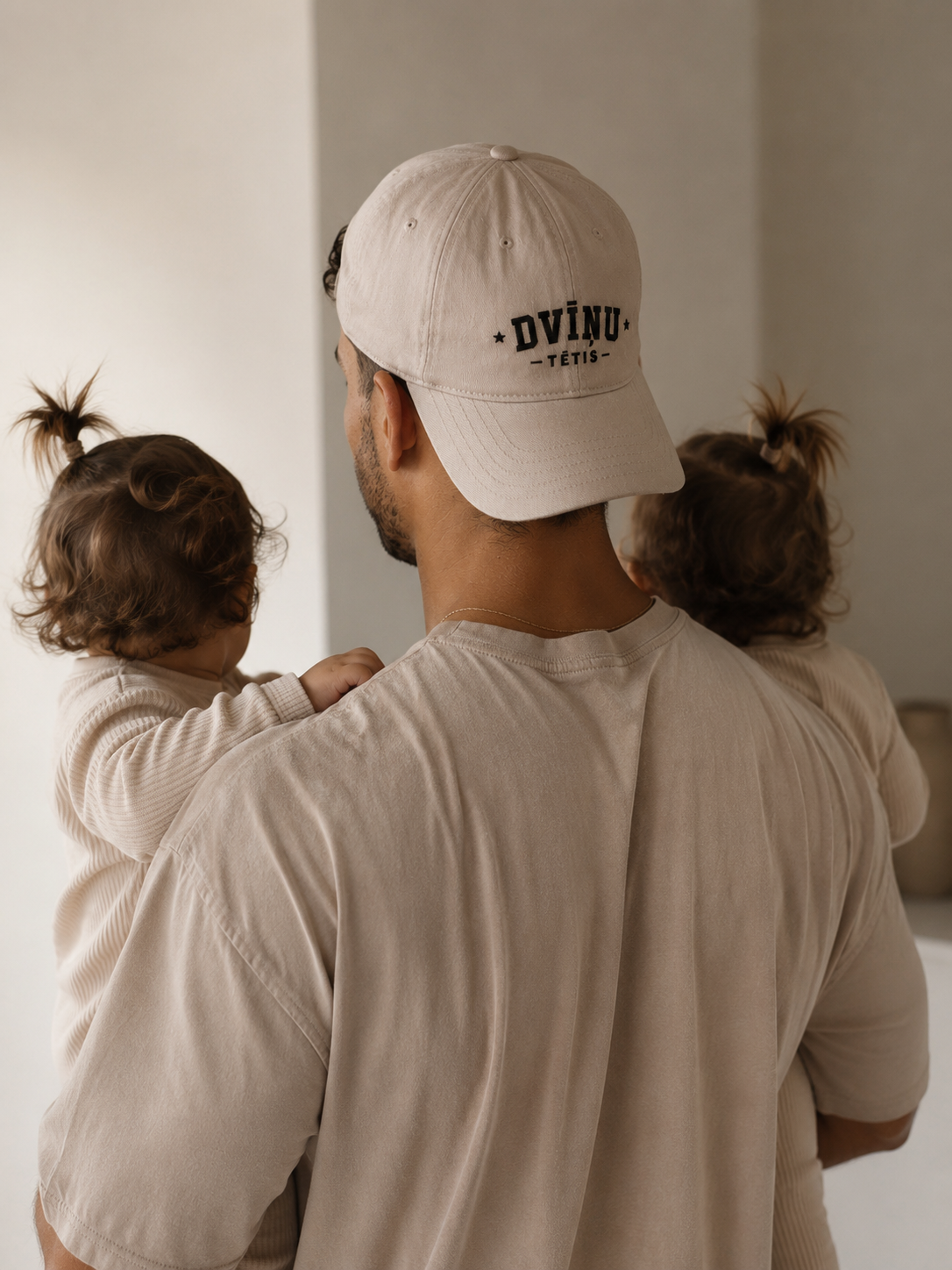 A man wearing a light beige baseball cap with black 'DVĪŅU tētis' text on the back, carrying two young children on his shoulders in a minimalist indoor setting.

