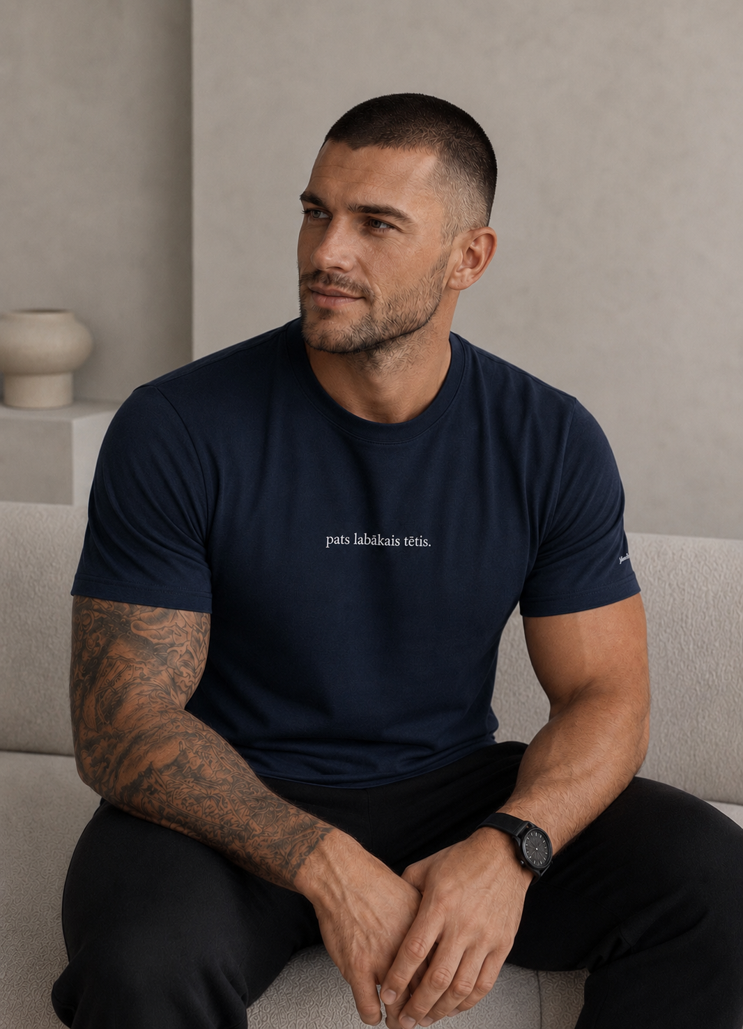 A man with a shaved head and tattoos sits on a light gray sofa wearing a navy blue t-shirt with white text on the chest that reads 'pats labākais tētis.'