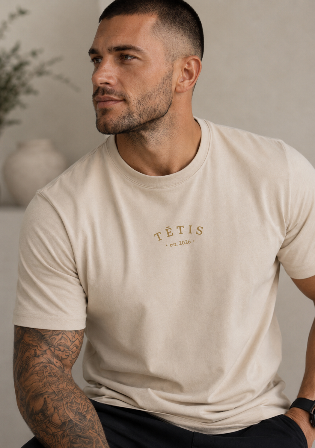 A man wearing a beige crew-neck t-shirt with 'TĒTIS EST. 2026' printed in gold on the chest, shown in a lifestyle setting.