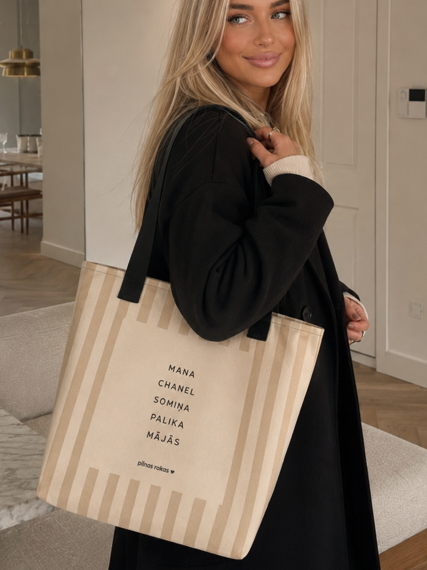 A woman in a black coat carries a beige canvas tote bag with vertical stripes and black text. The photo is taken in a modern, brightly lit interior with light-colored walls and wood floors.