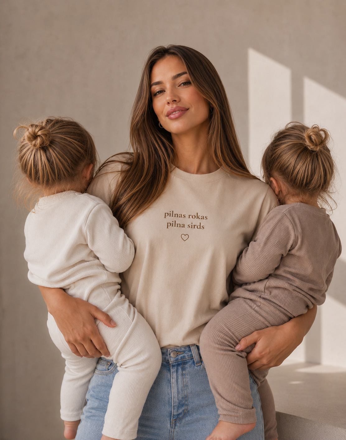 A woman holding two young children in a warm, minimalist room. She is wearing a beige crewneck t-shirt with the text 'pilnas rokas pilna sirds' and a small heart printed on the chest, paired with light-wash blue jeans. Brand of the t-shirt is "Pilnas Rokas"