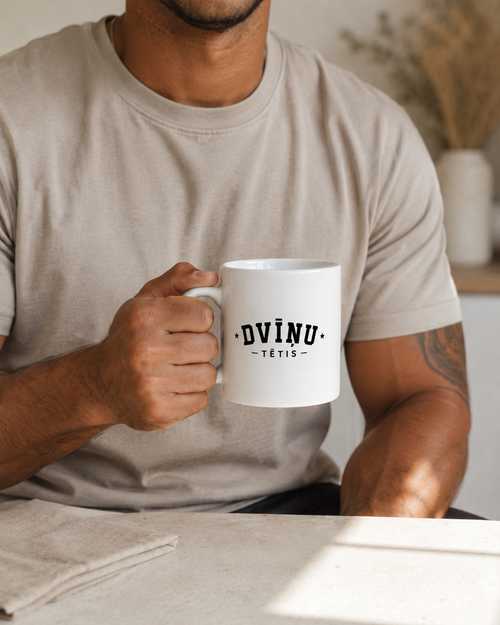 A man in a beige t-shirt holding a white ceramic mug with the text 'DVĪŅU TĒTIS ' printed in black.