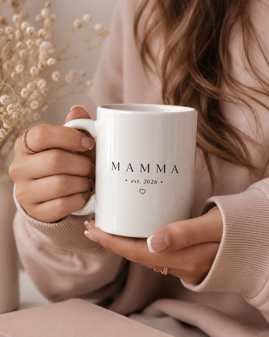 A person in a soft blush pink sweater holds a white ceramic mug with the word 'MAMMA' and small 'est. 2026' text printed in black.