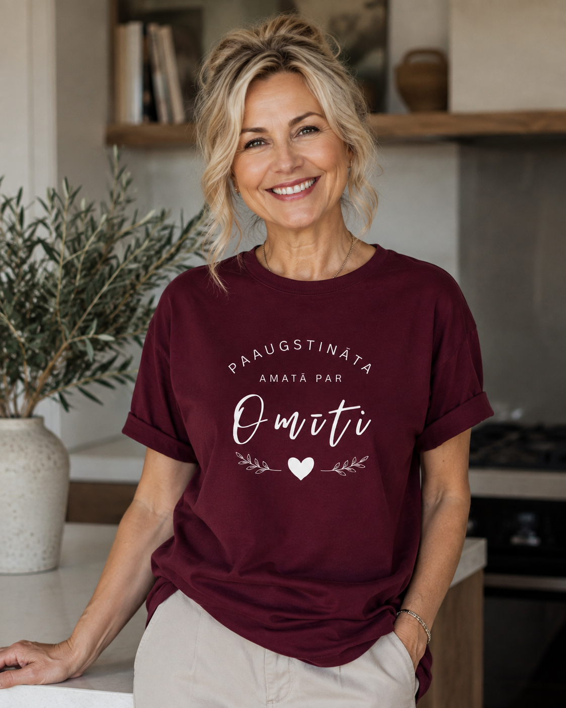 A smiling blonde woman wearing a maroon short-sleeve graphic t-shirt with white text that reads 'PAAUGSTINATA AMATA PAR OMĪTI' and a small heart, posed in a bright, modern kitchen setting.