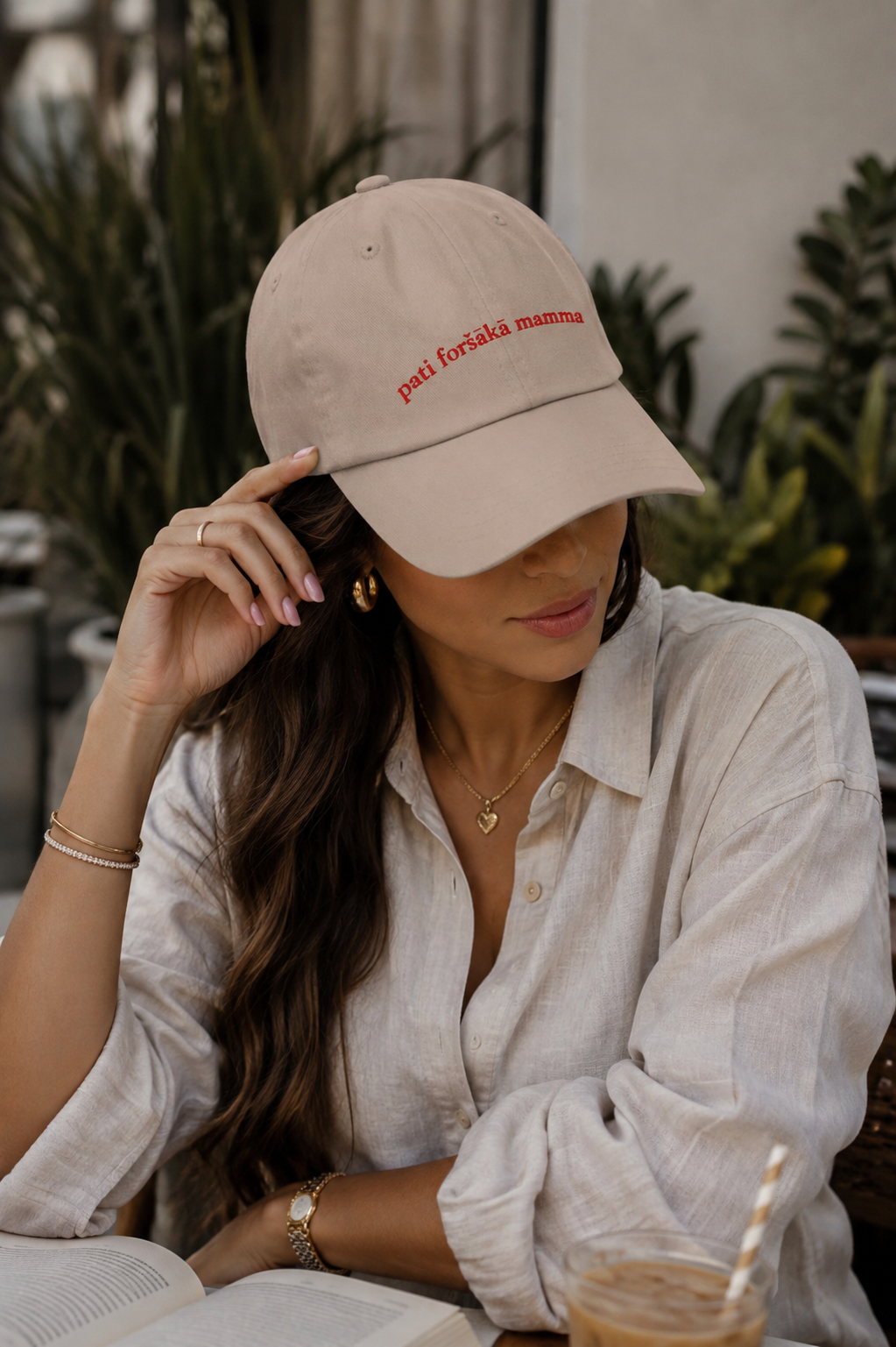 A woman wearing a beige baseball cap with red embroidered text reading 'pati foršākā mama' and a white linen shirt, seated at a table with an open book and an iced coffee in a cafe setting.