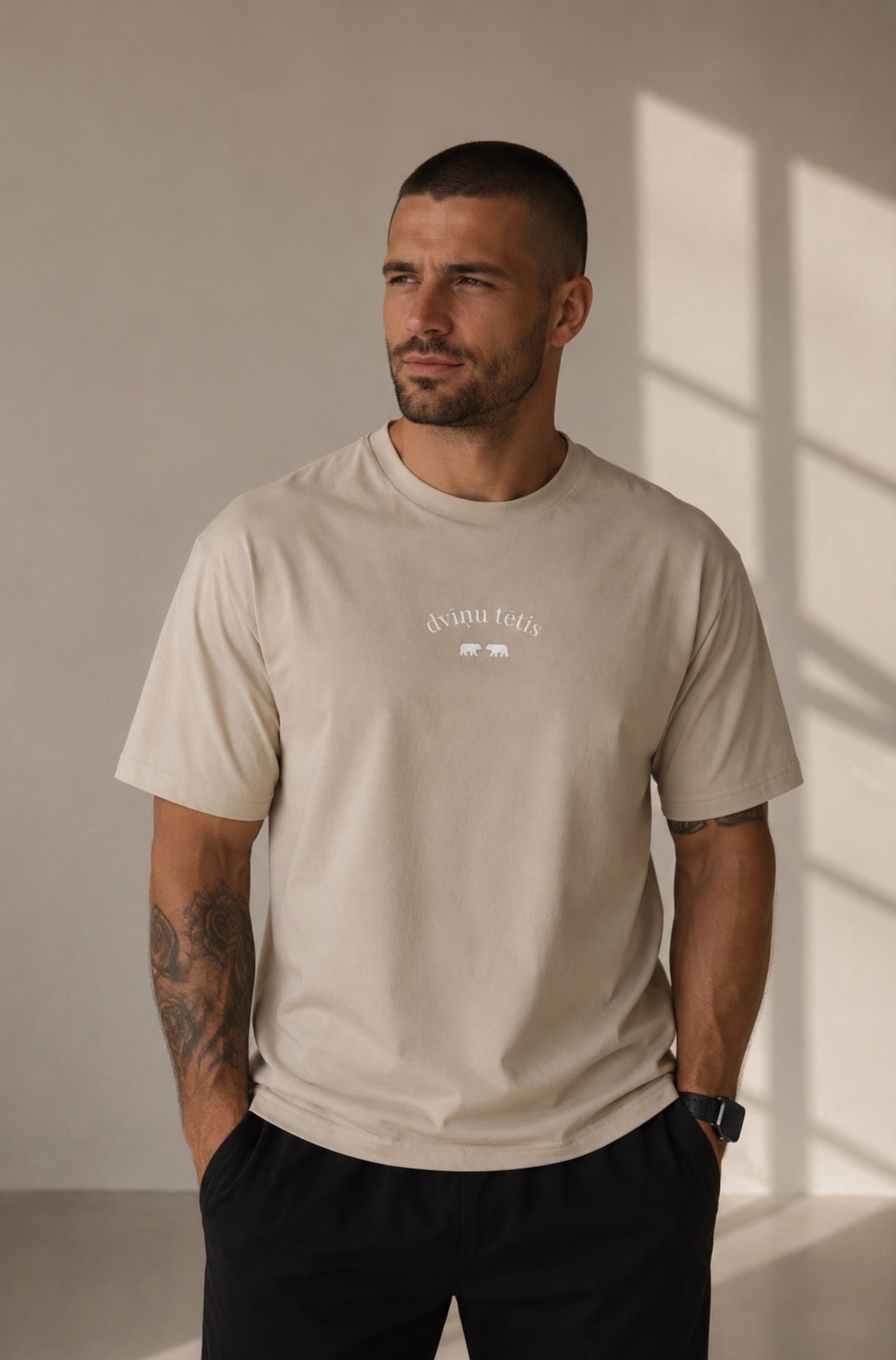 A man wearing a light beige crew-neck t-shirt with a small white graphic on the left chest, paired with black pants, standing in a brightly lit room with window shadows.