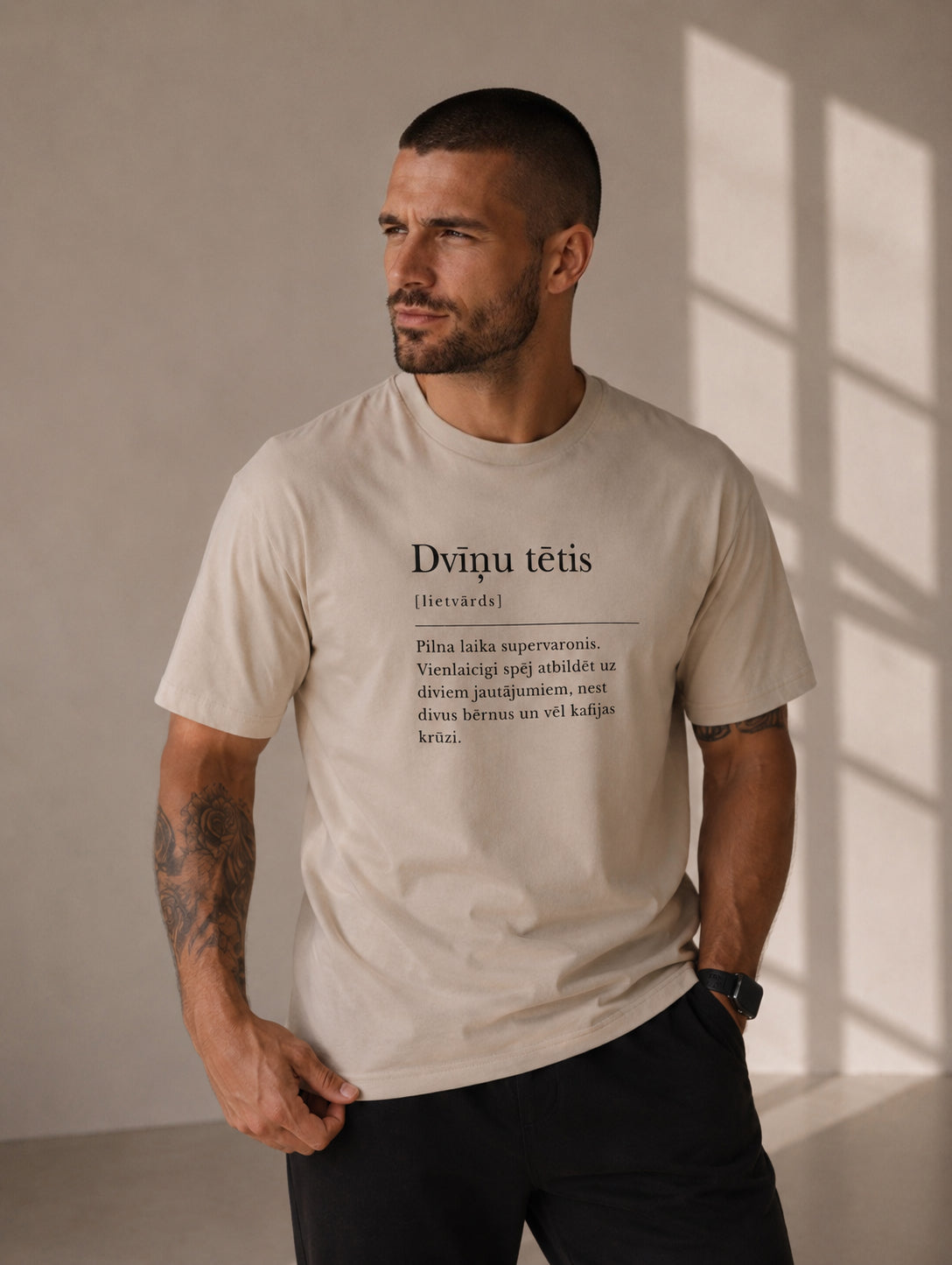 A man wearing a light beige graphic t-shirt with a humorous Latvian dictionary definition print of Dvīņu tētis, paired with black pants, standing in a room with warm window light casting shadows on the wall.