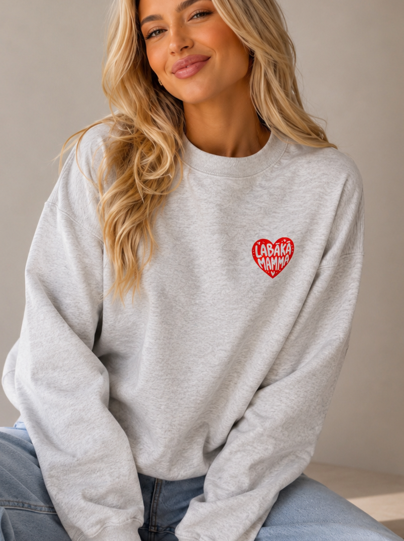 A woman with blonde hair sits wearing a light gray crewneck sweatshirt with a red heart graphic on the left chest that reads 'LABĀKĀ MAMMA', paired with light blue jeans against a neutral beige background.