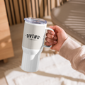 A hand holding a white insulated travel mug with a clear lid and black printed logo reading 'DVĪŅU TĒTIS' in a warm indoor setting.